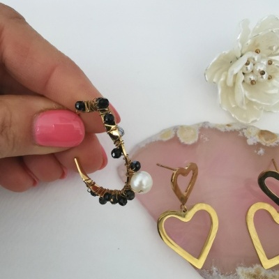 Brincos dourados com pedras pretas e pérola, coração dourado, sobre superfície branca e pedra rosa
