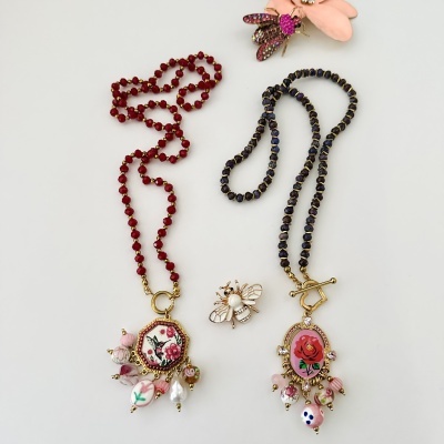 Dois colares com contas e pendentes florais, e um broche de abelha entre eles sobre fundo branco.