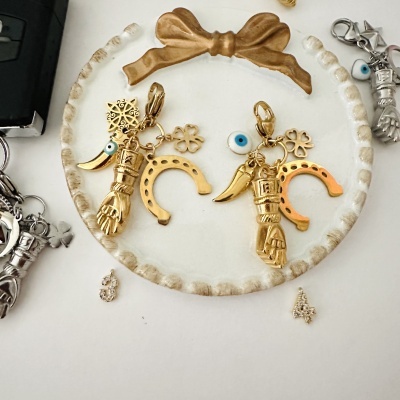 Par de chaveiros dourados com símbolos de mão, ferradura, trevo e olho azul sobre prato branco com laço dourado.