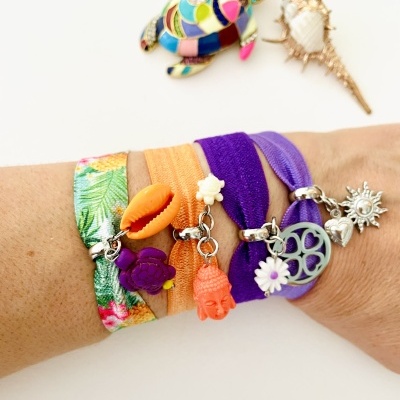 Braço com várias pulseiras de tecido coloridas com penduricalhos e dois broches coloridos no fundo.