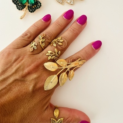 Mão com unhas rosa forte e anéis dourados de folhas, com dois broches de folhas ao fundo
