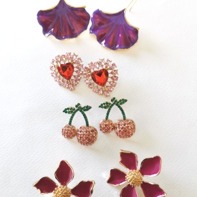 Cinco pares de brincos coloridos com flores, corações e cerejas sobre fundo branco.