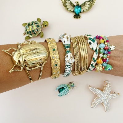 Pulseiras coloridas e douradas num braço com figuras decorativas de tartaruga, ave e estrela do mar ao fundo