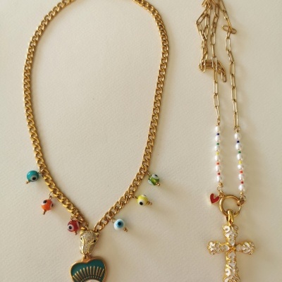 Dois colares dourados, um com pendentes coloridos de olho grego e um pingente em forma de coração com olho, outro com contas coloridas e cruz dourada.