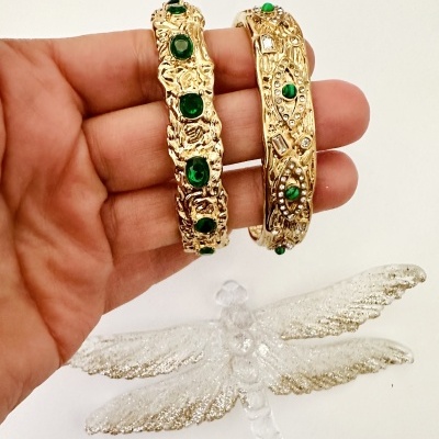 Duas pulseiras douradas com pedras verdes na mão sobre fundo branco com objeto prateado em forma de libélula