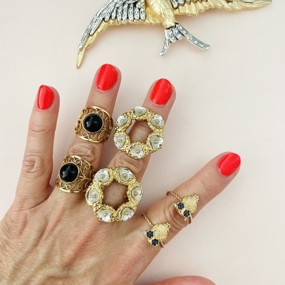 Mão com várias argolas douradas e pedras pretas e brancas, unhas vermelhas, broche de pássaro dourado e prateado