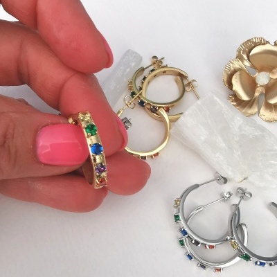 Brincos de argola dourados e prateados com pedras coloridas sobre fundo branco, mão segurando brinco dourado.