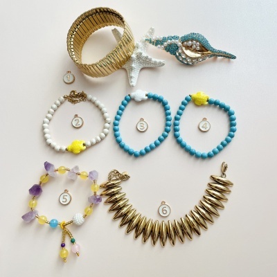 Pulseiras e broches coloridos, incluindo dourado, azul, branco e roxo, dispostos em fundo branco.