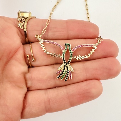 Colar dourado com pendente de pássaro colorido e anel dourado com pedras exibidos numa mão