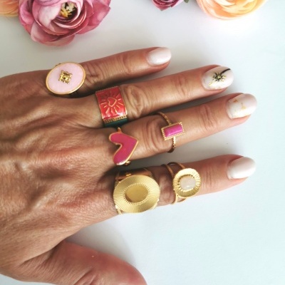 Mão com vários anéis dourados e vermelhos e unhas decoradas, flores ao fundo