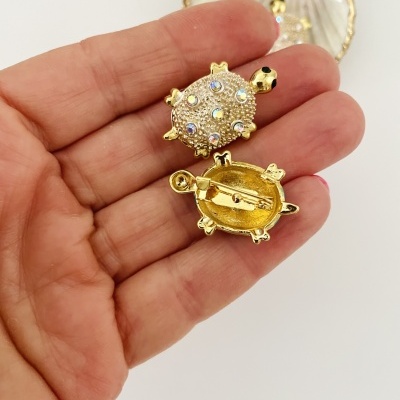 Broche de tartaruga dourado com pedras brilhantes na mão, com concha decorativa no fundo