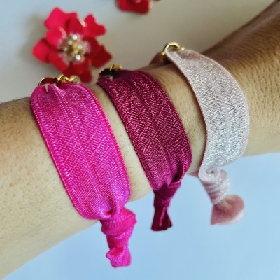 Três pulseiras elásticas de tecido brilhante em tons de rosa no pulso com flores decorativas ao fundo