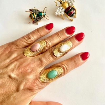 Mão com três anéis dourados com pedras coloridas e duas broches de abelhas douradas com pérolas no fundo