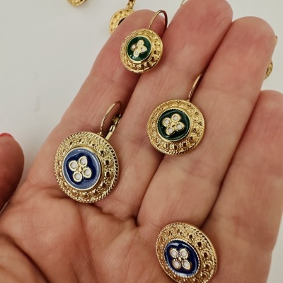 Brincos dourados redondos com detalhes em esmalte azul e verde, segurados numa mão.