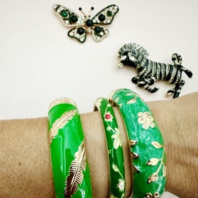 Três pulseiras verdes com padrões dourados e dois broches de borboleta e zebra sobre fundo claro.