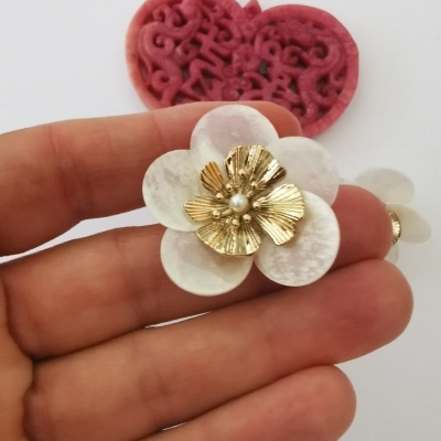Brinco flor branco com centro dourado e pérola segurado por mão com fundo coração vermelho