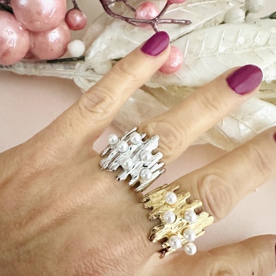 Dois anéis metálicos, um dourado e outro prateado, com pérolas brancas e unhas pintadas de vermelho escuro.