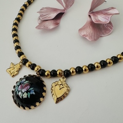 Colar com contas pretas e douradas e três pendentes sobre fundo branco com flores cor-de-rosa