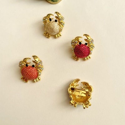 Broches em forma de caranguejo dourados com corpo colorido e detalhes brilhantes, sobre fundo claro com concha