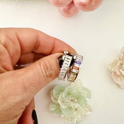 Brincos argola com pedras coloridas e brancas segurados por mão com unhas pintadas de preto