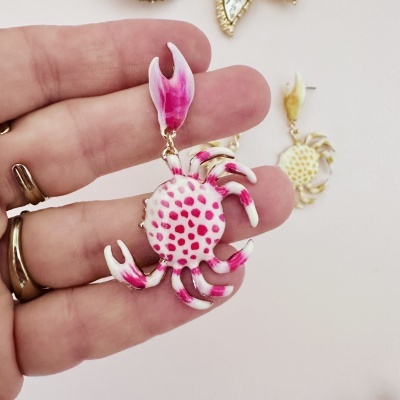Brinco em forma de caranguejo branco com manchas rosa sendo segurado por dedos, com outro brinco ao fundo.
