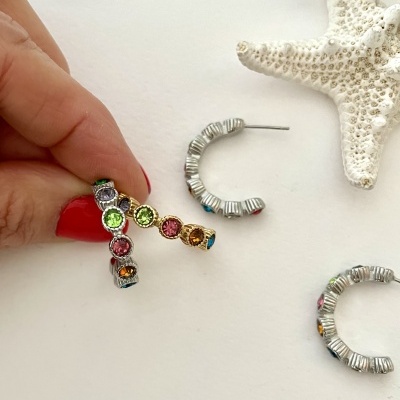 Brincos de argola em metal prateado com pedras coloridas, uma mão segurando um brinco sobre fundo branco com estrela do mar