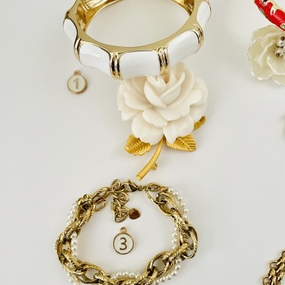 Tres pulseiras de metal dourado, uma com detalhes brancos e outra com pérolas, com etiquetas numeradas e uma flor branca decorativa.