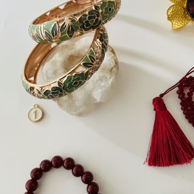 Braceletes douradas com padrão floral verde e branco sobre pedra branca e pulseiras coloridas ao lado.
