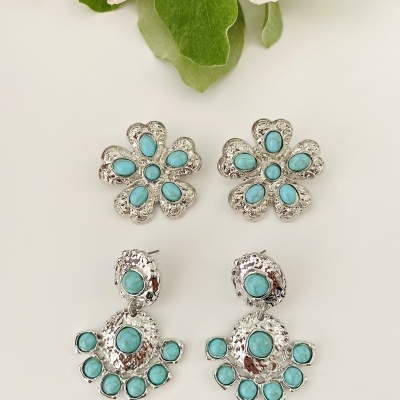 Dois pares de brincos prateados com pedras turquesa em forma de flor e meia-lua sobre fundo branco com flores ao fundo.