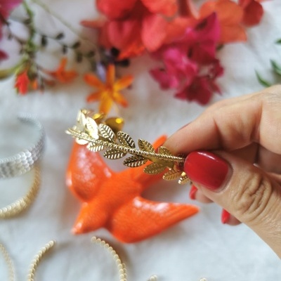 Presilha dourada em forma de folhas segurada por mão com unhas vermelhas