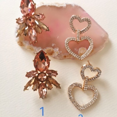 Dois pares de brincos, um floral castanho-dourado e outro de corações dourados com cristais, sobre fundo branco e pedra rosa