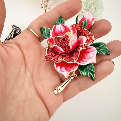 Broche de rosa vermelha com pedras vermelhas e folhas verdes numa mão.