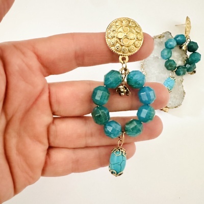 Brincos dourados com pedras azuis em argola e pendente azul sendo segurados por uma mão