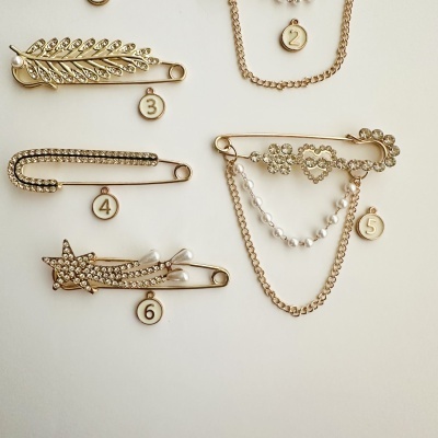 Alfinetes dourados decorativos com pedras transparentes e pérolas, cada um com etiqueta numérica, em fundo branco.