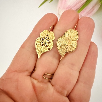 Brincos dourados com padrão floral, um com detalhes azuis, na mão com fundo branco e flores rosas