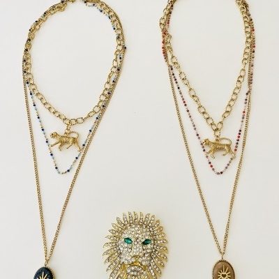 Dois colares dourados com pingentes de elefante e broche de cabeça de leão sobre fundo branco