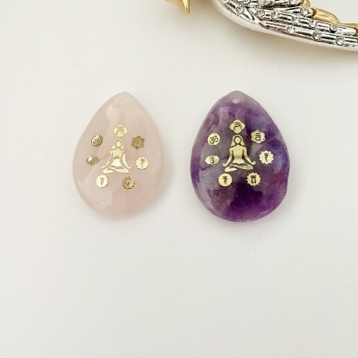 Duas pedras em formato de gota com símbolos dourados, uma rosa e outra roxa, sobre fundo branco com objeto decorativo prateado e dourado