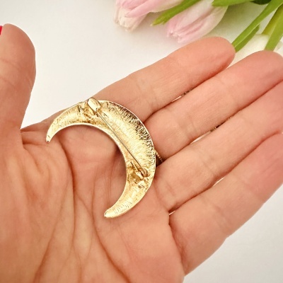 Broche dourado em forma de lua crescente numa mão com flores ao fundo