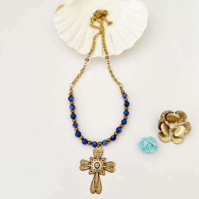 Colar dourado com contas azuis e pendente cruz, sobre concha branca, com flores decorativas ao lado.