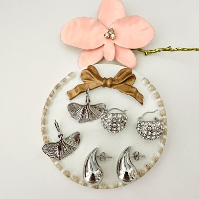 Três pares de brincos prateados em prato branco com laço castanho e flor rosa ao fundo
