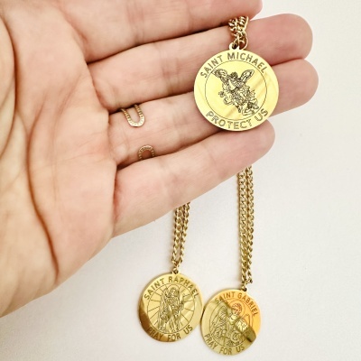 Colares dourados com medalhões redondos e textos de proteção em inglês, apresentados numa mão.