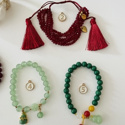 Três pulseiras de contas numeradas em fundo branco, nas cores vermelho, verde claro e verde escuro, com pendentes e detalhes dourados.