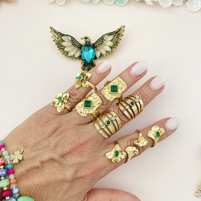 Mão com vários anéis dourados com pedras verdes e pulseiras coloridas