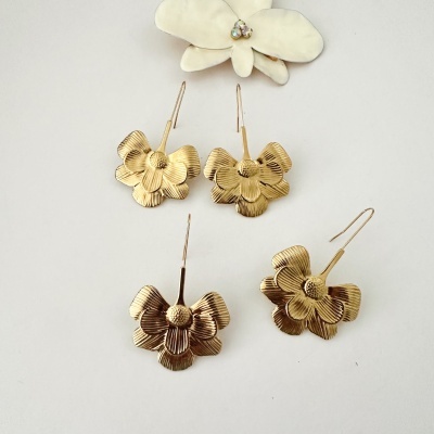 Brincos dourados em forma de flor com textura, sobre superfície branca com flor decorativa ao fundo