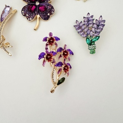 Broches decorativos metálicos com pedras em formas florais e de folhas, cores roxo, verde e dourado, fundo branco.