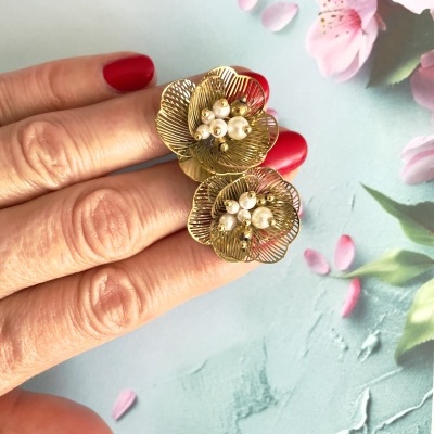 Anel dourado de flores com pérolas em dedo com unhas vermelhas sobre fundo azul claro com flores cor-de-rosa