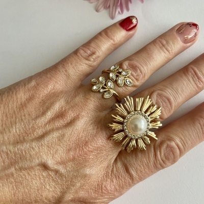 Mão com unhas decoradas e dois anéis dourados, um com pedras claras e outro com pérola, sobre fundo branco com flores