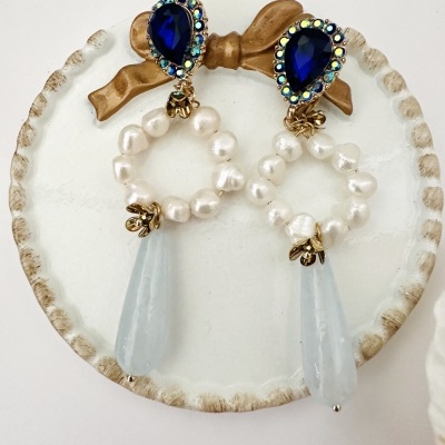 Brincos com pedras azuis, pérolas e pendente azul claros num prato cerâmico branco com laço castanho.
