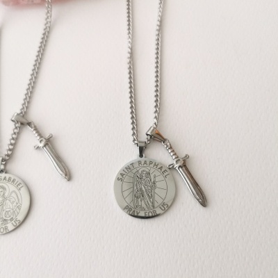 Dois colares prateados com pendentes redondos religiosos e uma espada pequena, sobre superfície branca com pedra rosa.