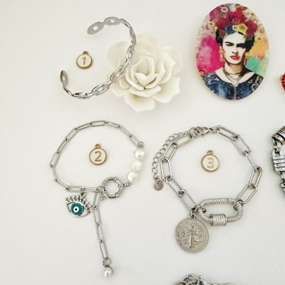 Três pulseiras prateadas com diferentes pendentes numeradas 1 a 3, sobre fundo branco com flor e imã colorido.